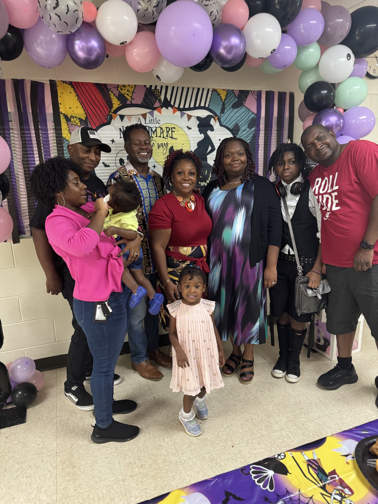 Large family group gathered at a festive event under purple and pink balloons, with Ivy’s mother in the center smiling among children and loved ones.